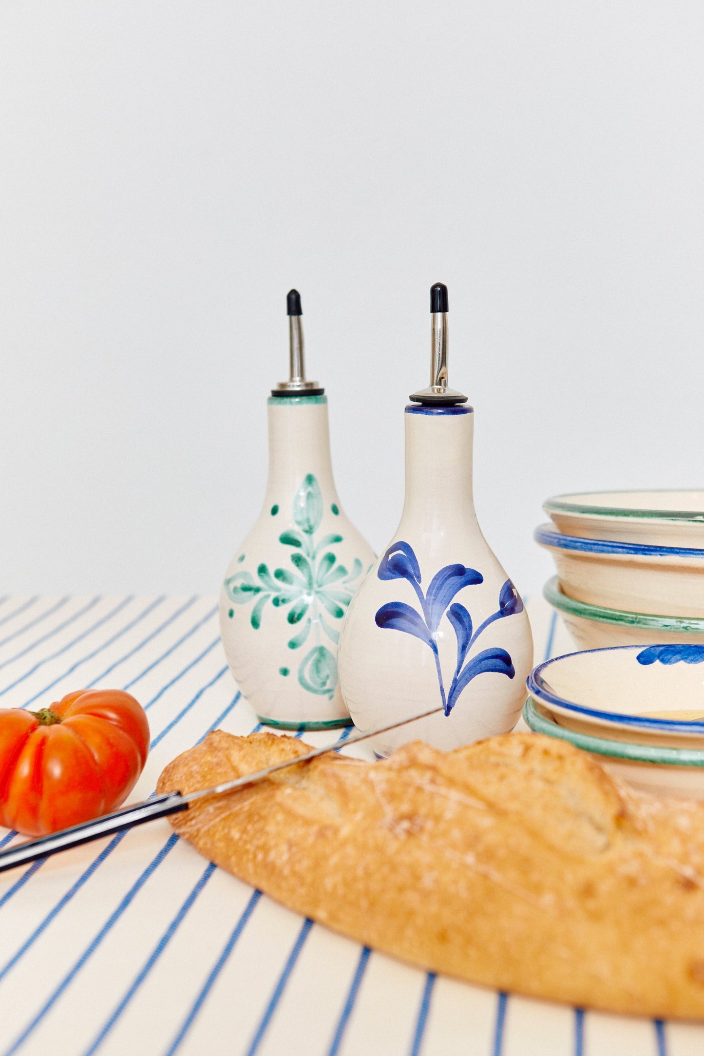 Juego de aceiteras de cerámica blanca y azul, junto a un tomate y pan fresco sobre un mantel a rayas.