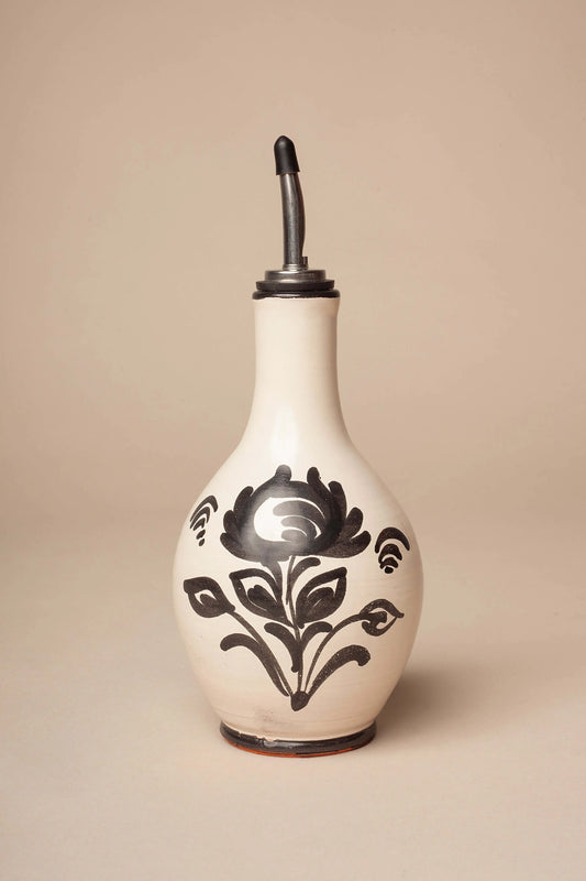 Aceitera de cerámica crema con forma de pera y dibujo floral en negro, rematada con un vertedor de metal.