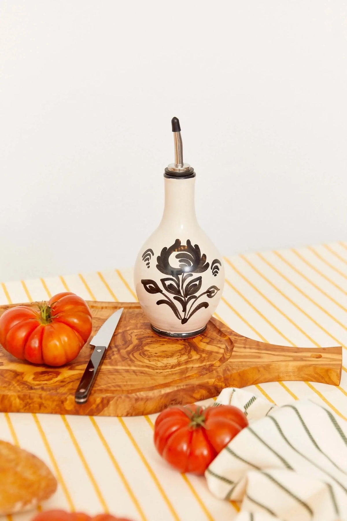 Aceitera de cerámica crema con motivos florales negros, tapón metálico, sobre tabla de madera con tomates y cuchillo.