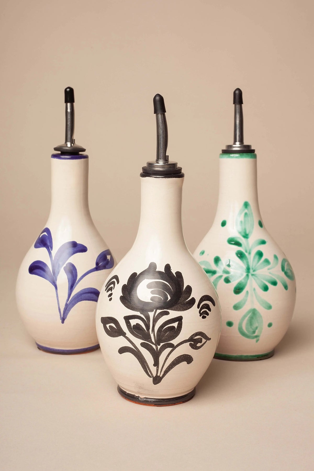 Tres aceiteras de cerámica blanca con diseños florales pintados a mano en azul, negro y verde, y vertedor metálico.