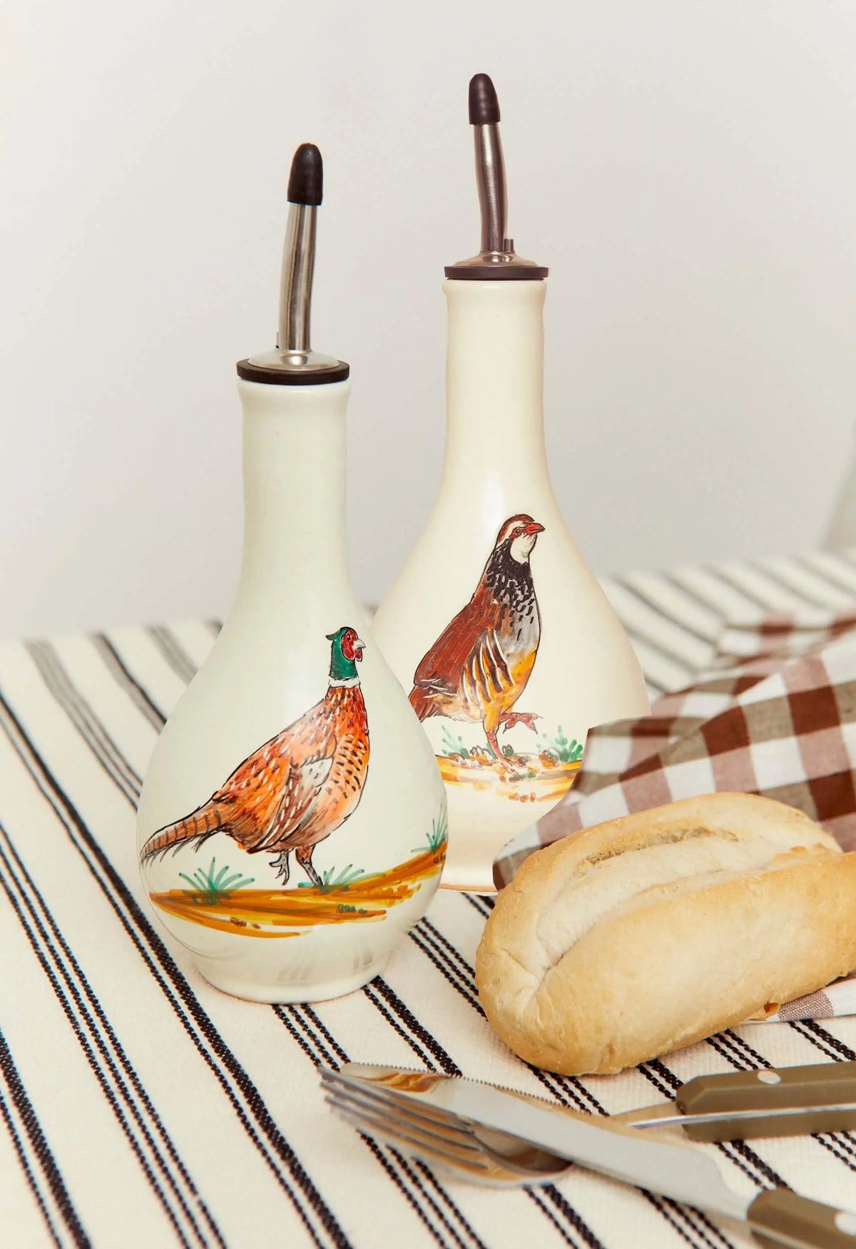 Dos aceiteras de cerámica blanca decoradas con aves, estilo artesanal, sobre mantel a rayas con pan y cubiertos.