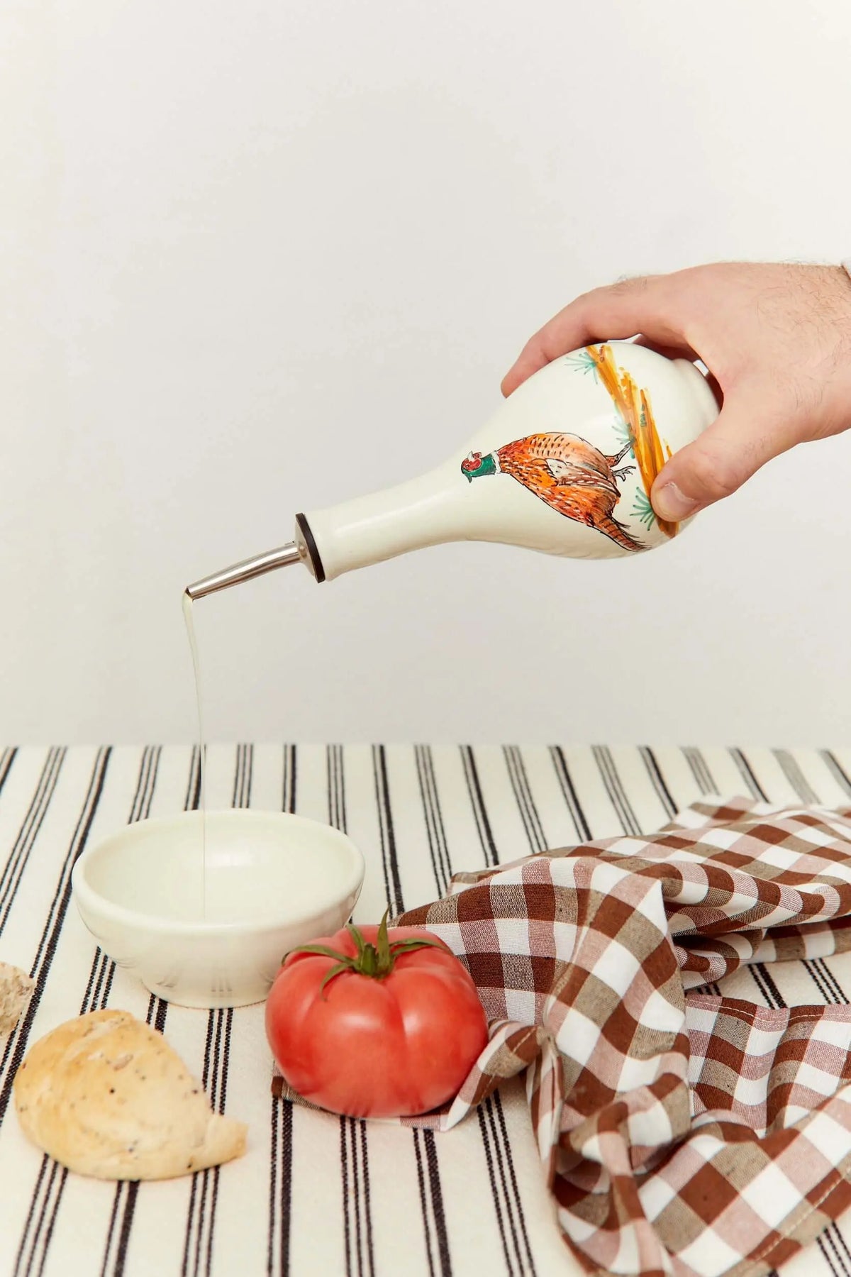 Aceitera de cerámica con estampado de faisán sirviendo aceite sobre un cuenco, junto a un tomate y un paño a cuadros.