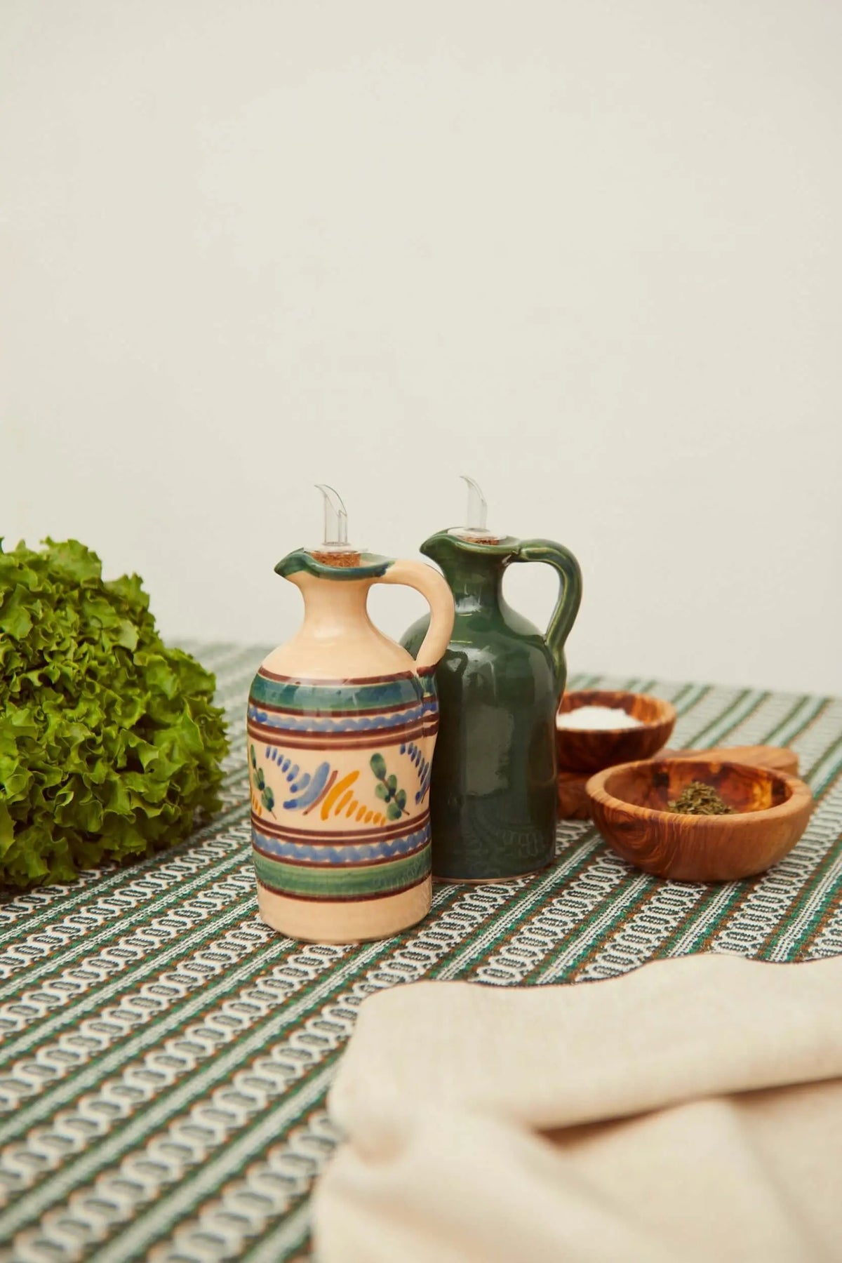 Dos aceiteras de cerámica artesanal, una decorada con motivos geométricos y otra verde liso, sobre un mantel verde.