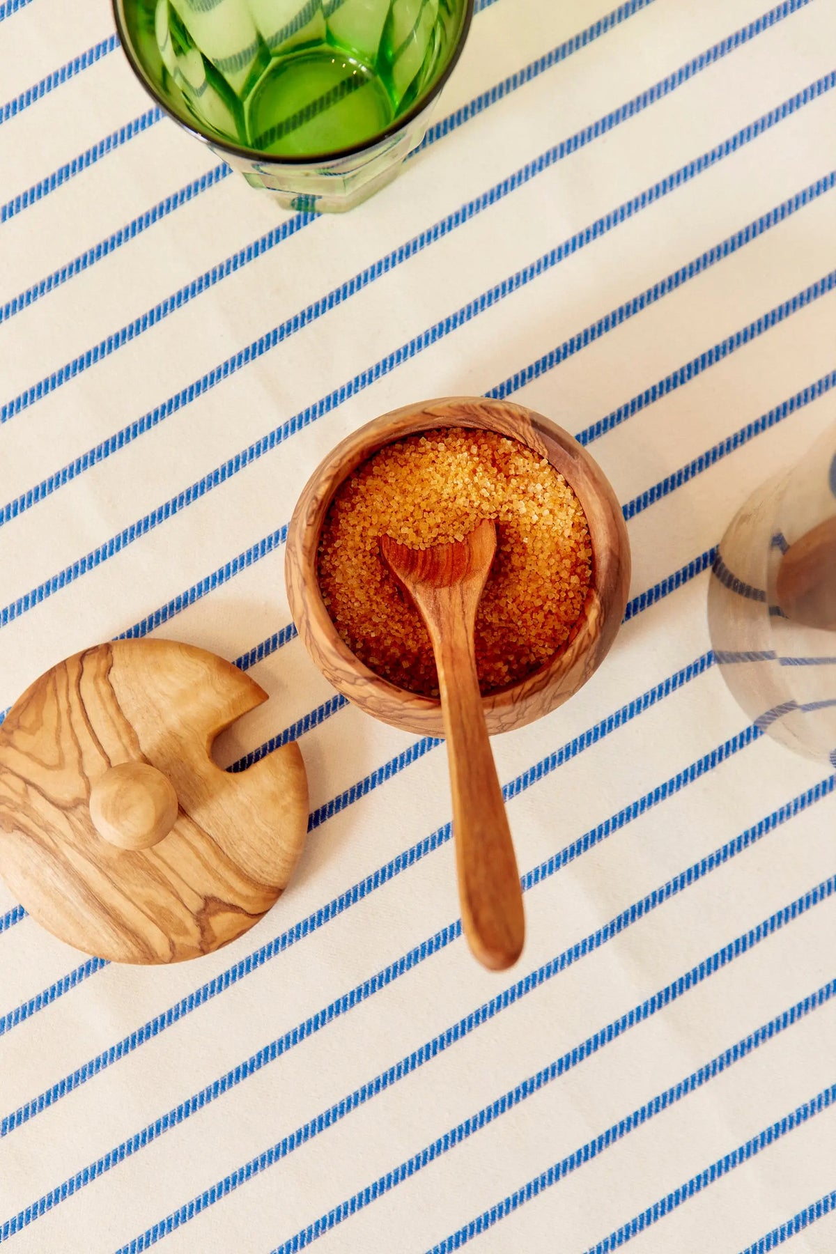 Azucarero de madera de olivo con tapa y cucharilla, lleno de azúcar moreno sobre un mantel de rayas azules y blancas.
