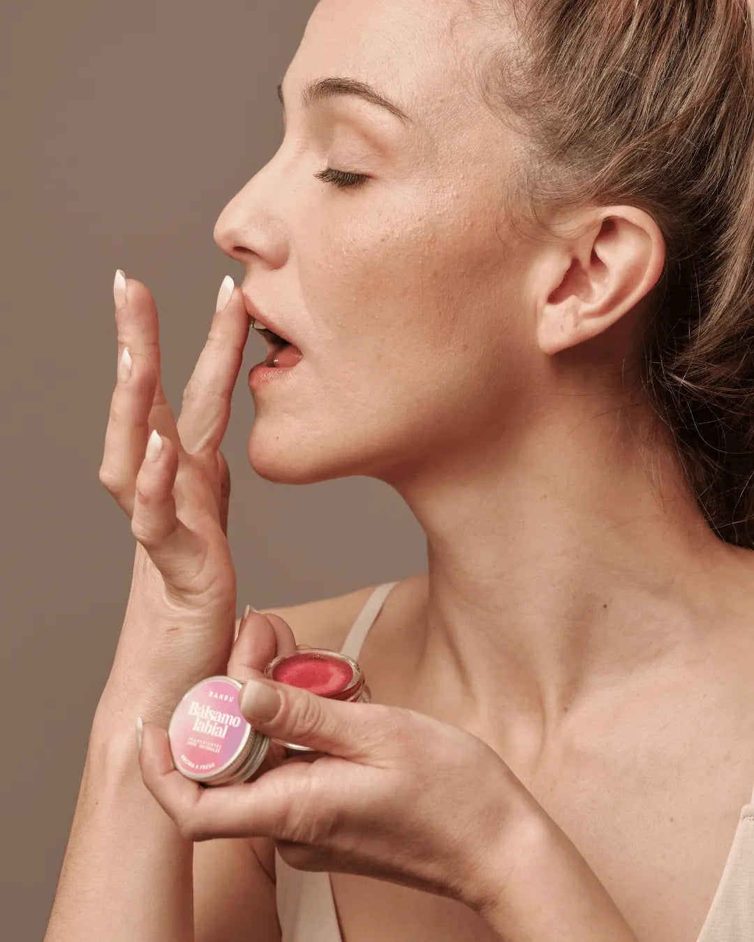 Mujer aplicando bálsamo labial Banbu de color rojo, extraído de un pequeño tarro de aluminio sobre fondo neutro.