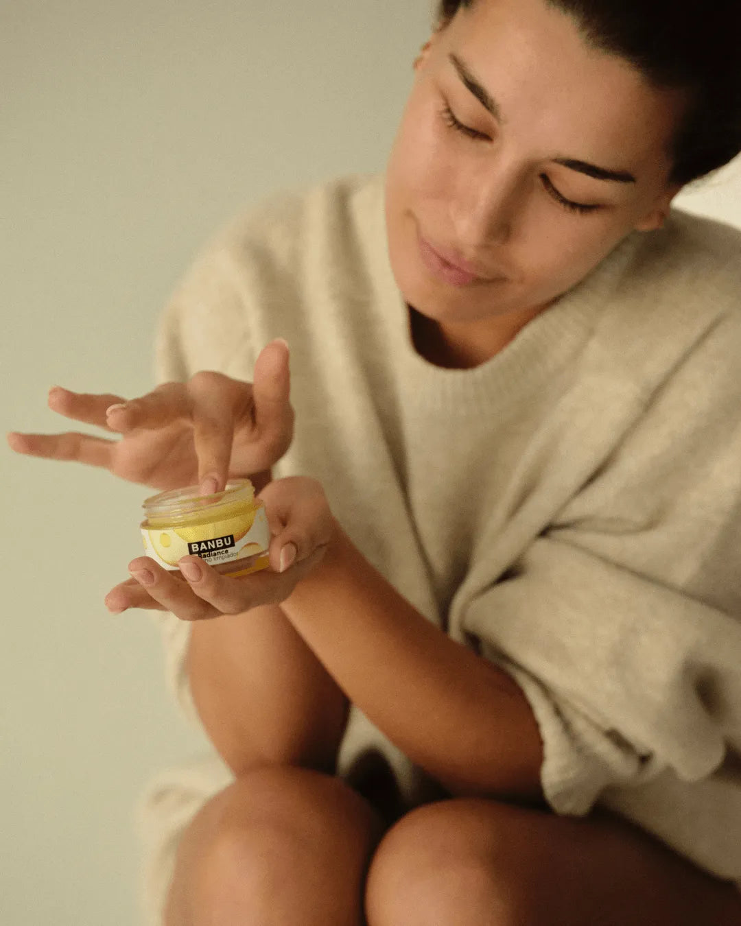Mujer aplica bálsamo limpiador facial Radiance de Banbu, de textura cremosa y envase amarillo, sobre un fondo neutro.