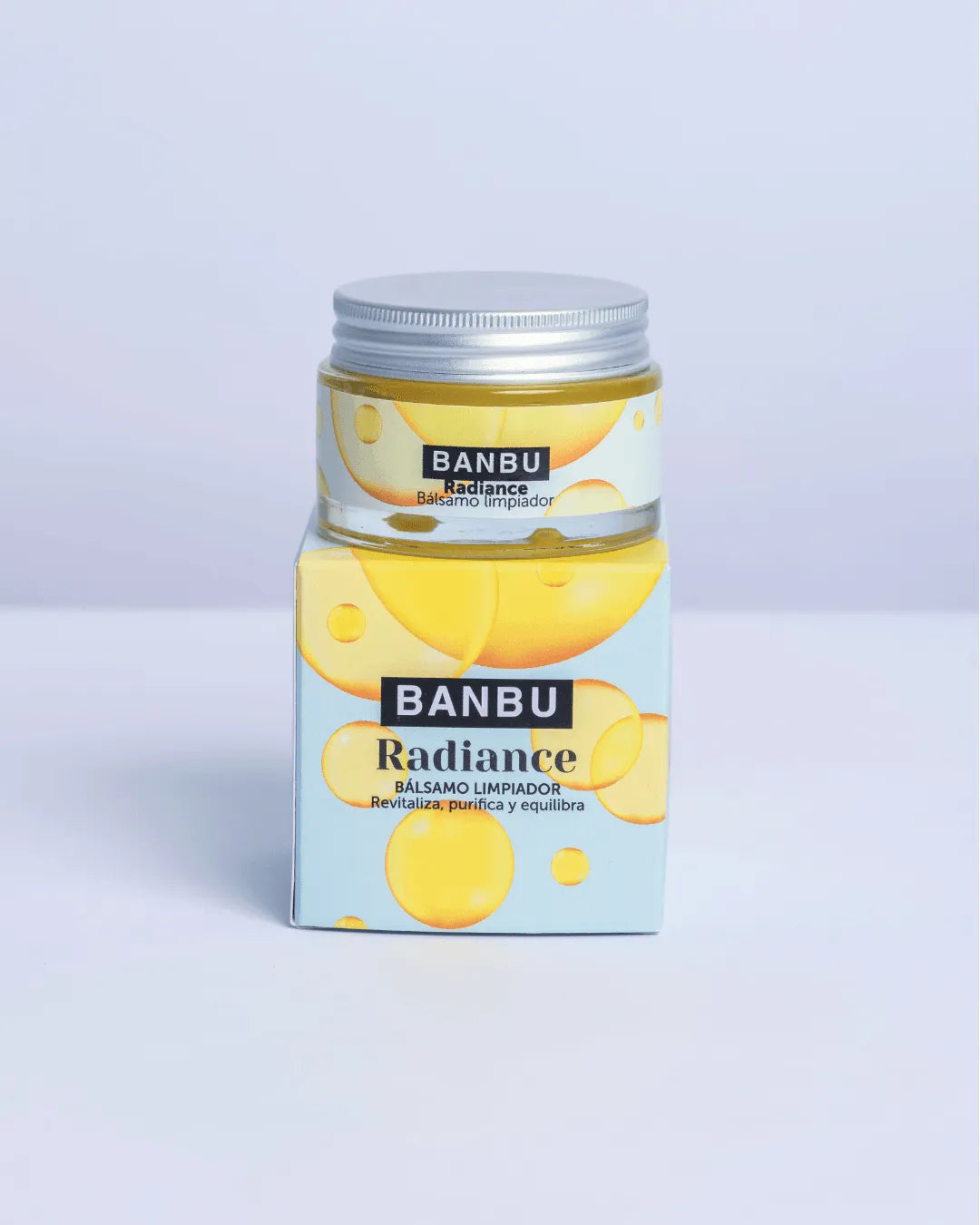 Bálsamo limpiador Banbu Radiance en tarro de vidrio sobre su caja azul con burbujas amarillas. Fondo blanco.