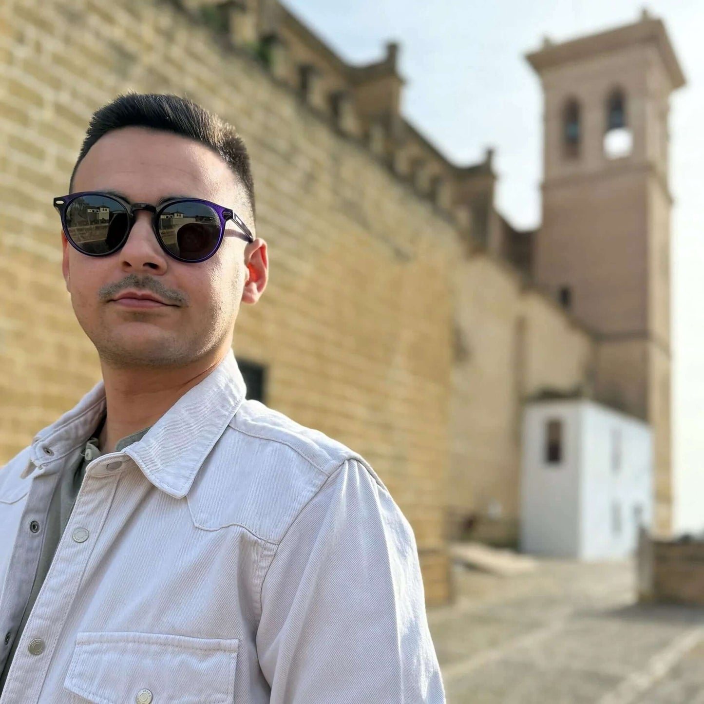 Hombre usando gafas de sol redondas con montura color púrpura y lentes oscuros, con una arquitectura histórica de fondo.