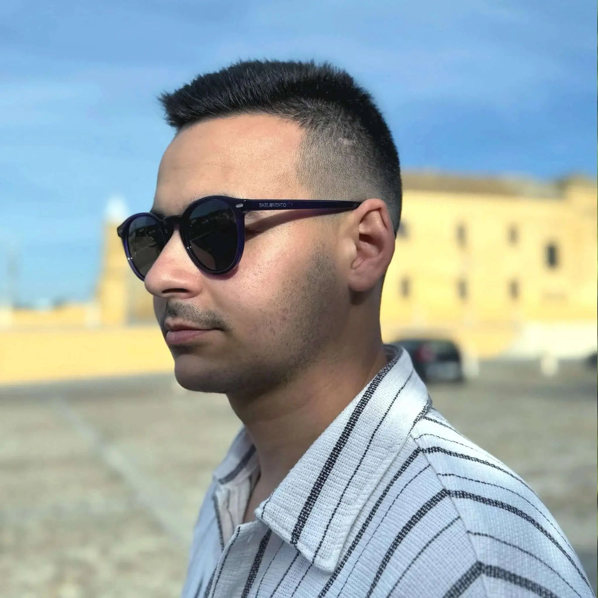 Gafas de sol de montura redonda en color morado oscuro con lentes oscuras puestas por un hombre de perfil.