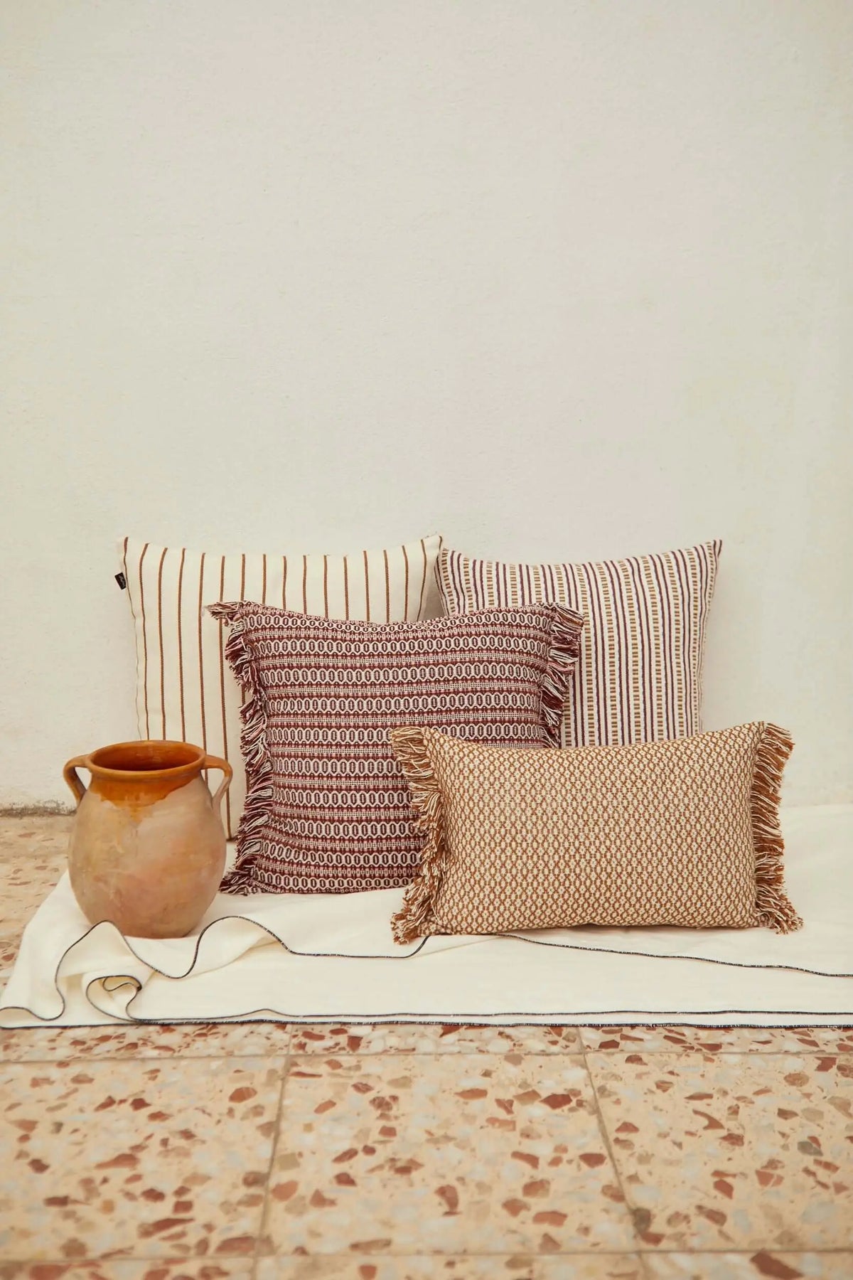 Four decorative cushions with stripes, geometric patterns, and fringed edges in maroon, beige, and white on a floor.