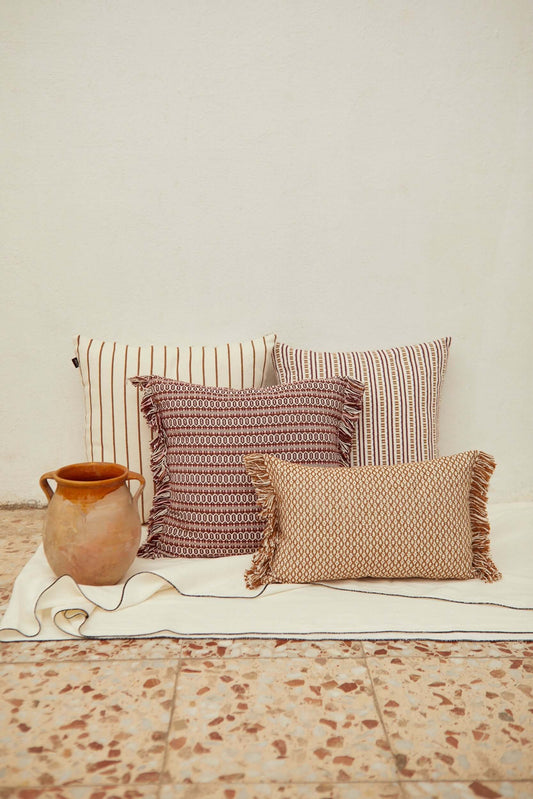 Four decorative cushions with stripes, geometric patterns, and fringed edges in maroon, beige, and white on a floor.