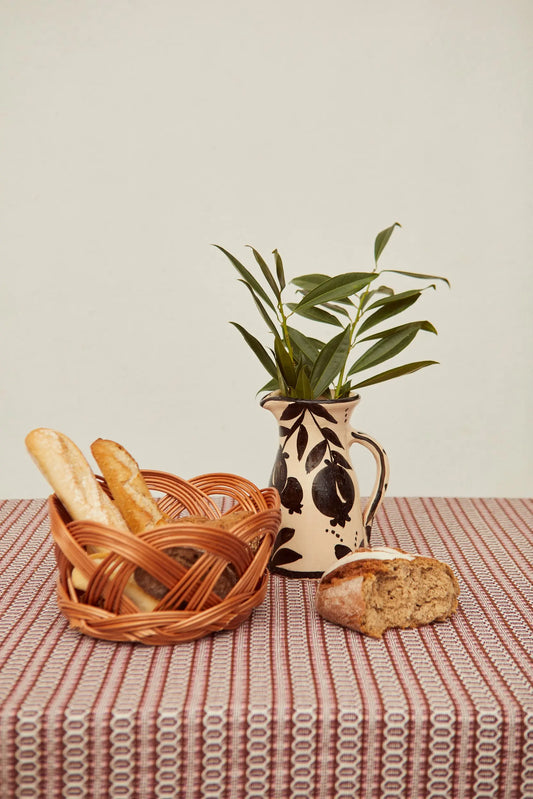 Panera redonda de mimbre natural con diseño calado, sobre mantel de rayas, junto a una jarra decorativa y pan artesano.