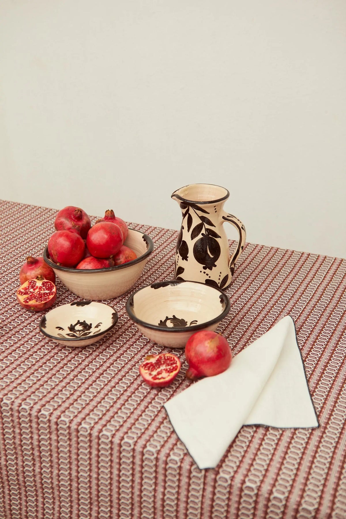 Set of cream-colored ceramic bowls with black pomegranate illustrations, placed on a patterned tablecloth with fruit.