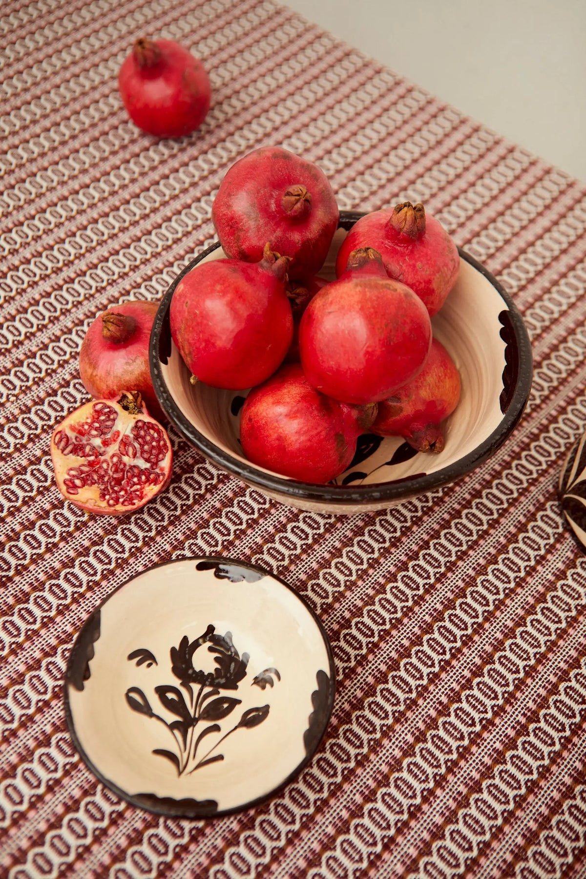 Handcrafted ceramic bowls in cream with black floral patterns, filled with fresh pomegranates on a textured cloth.