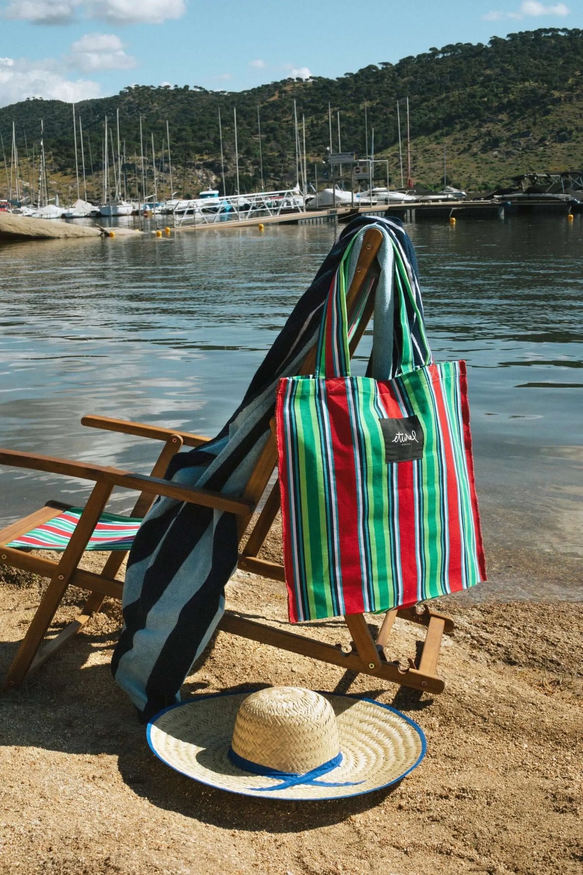 Hamaca de madera con tejido de lona a rayas rojas y verdes, una bolsa a juego y una toalla sobre un fondo de puerto.