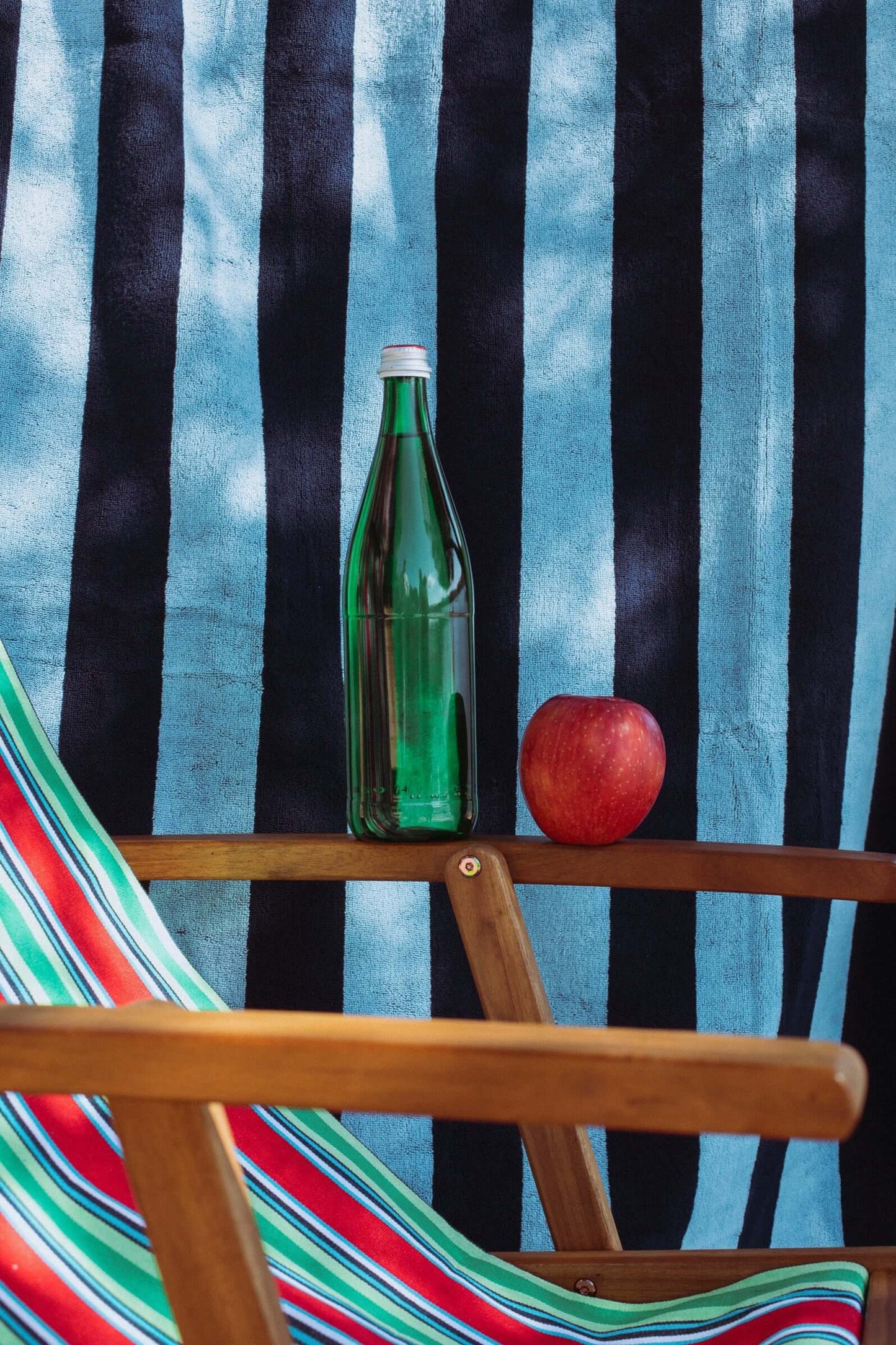 Tejido a rayas rojas, verdes y blancas en hamaca de madera, con una botella de vidrio y una manzana roja.
