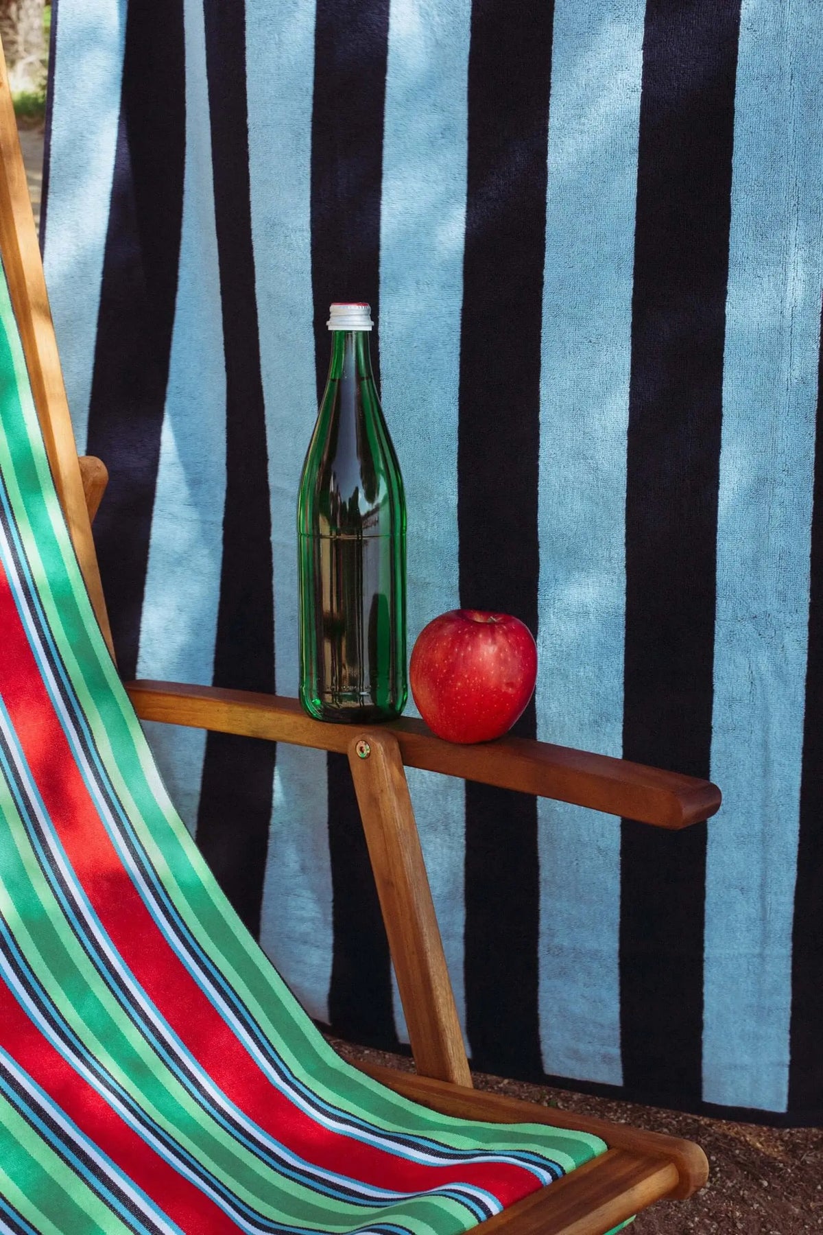 Navy blue and light blue striped cotton beach towel draped behind a wooden deck chair with a bottle and an apple.
