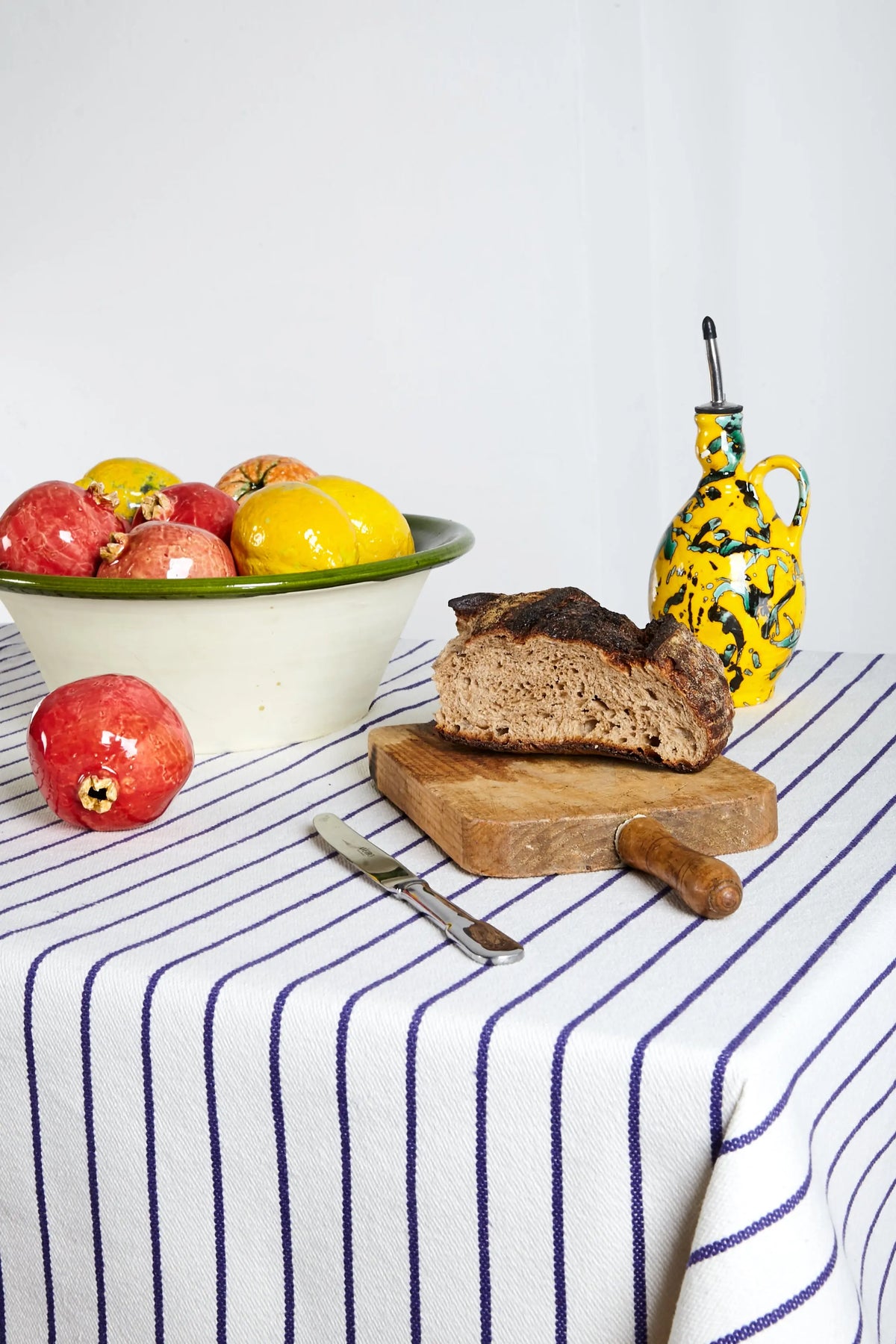 Pan marrón sobre una tabla de madera, con bowl de frutas coloridas y jarra amarilla decorativa, sobre manteles a rayas.