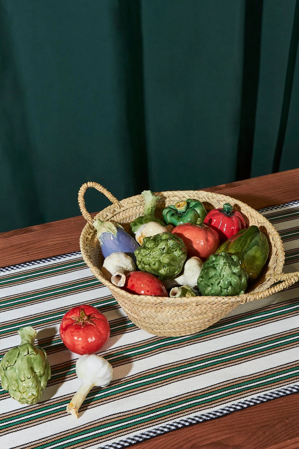 Cesta de mimbre con verduras de cerámica en colores rojo, verde y morado sobre un mantel a rayas.