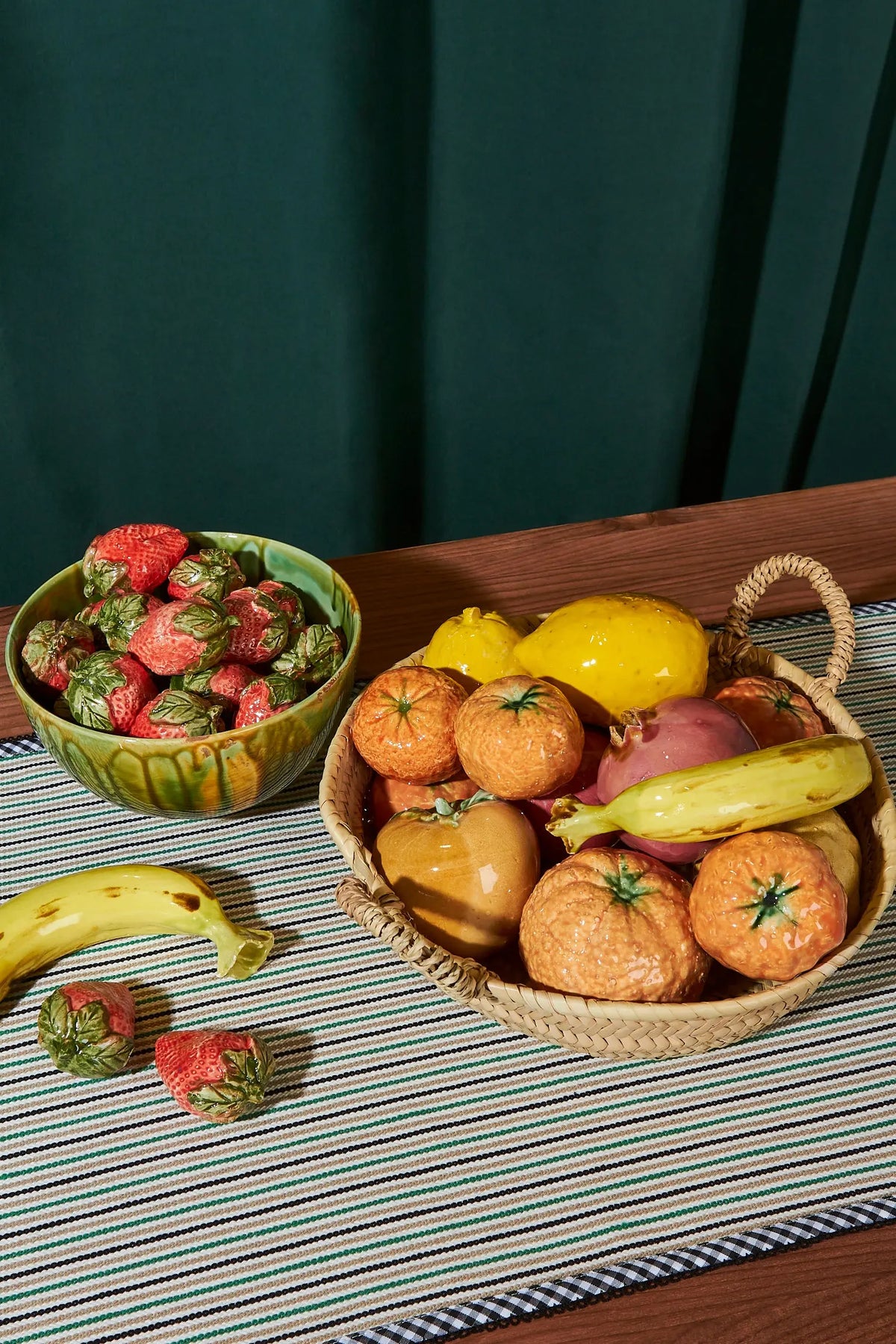 Frutas de cerámica en tonos naranja y amarillo en canasta de mimbre, con fresas en tazón verde en mesa de rayas.
