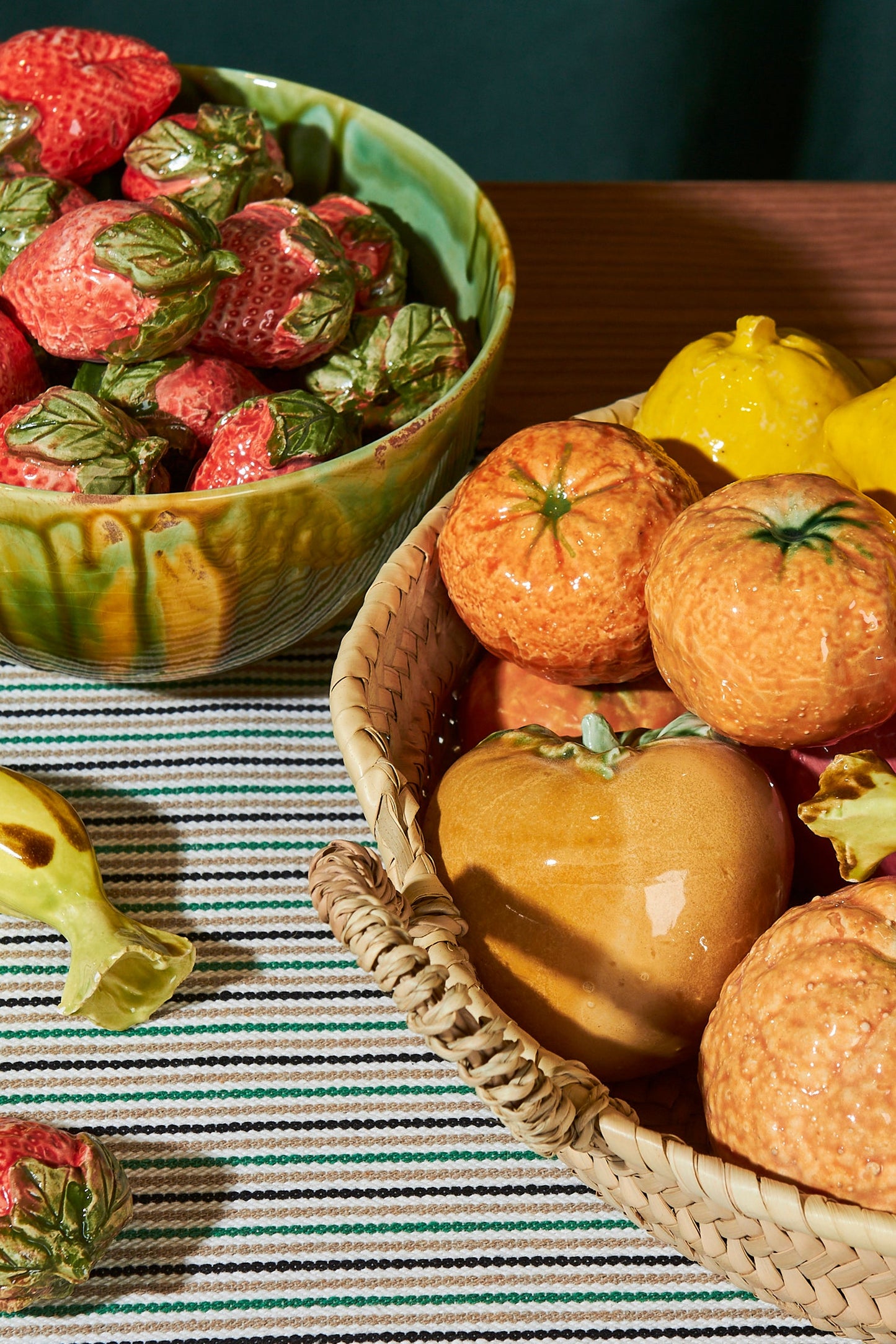 Frutas decorativas de cerámica en tonos naranja y amarillo, en cesto de mimbre y tazón de cerámica verde.