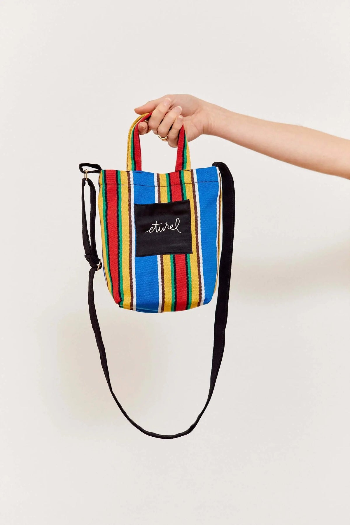 Bolso bandolera mini de lona a rayas multicolor con asas cortas y correa negra ajustable. Estilo casual veraniego.