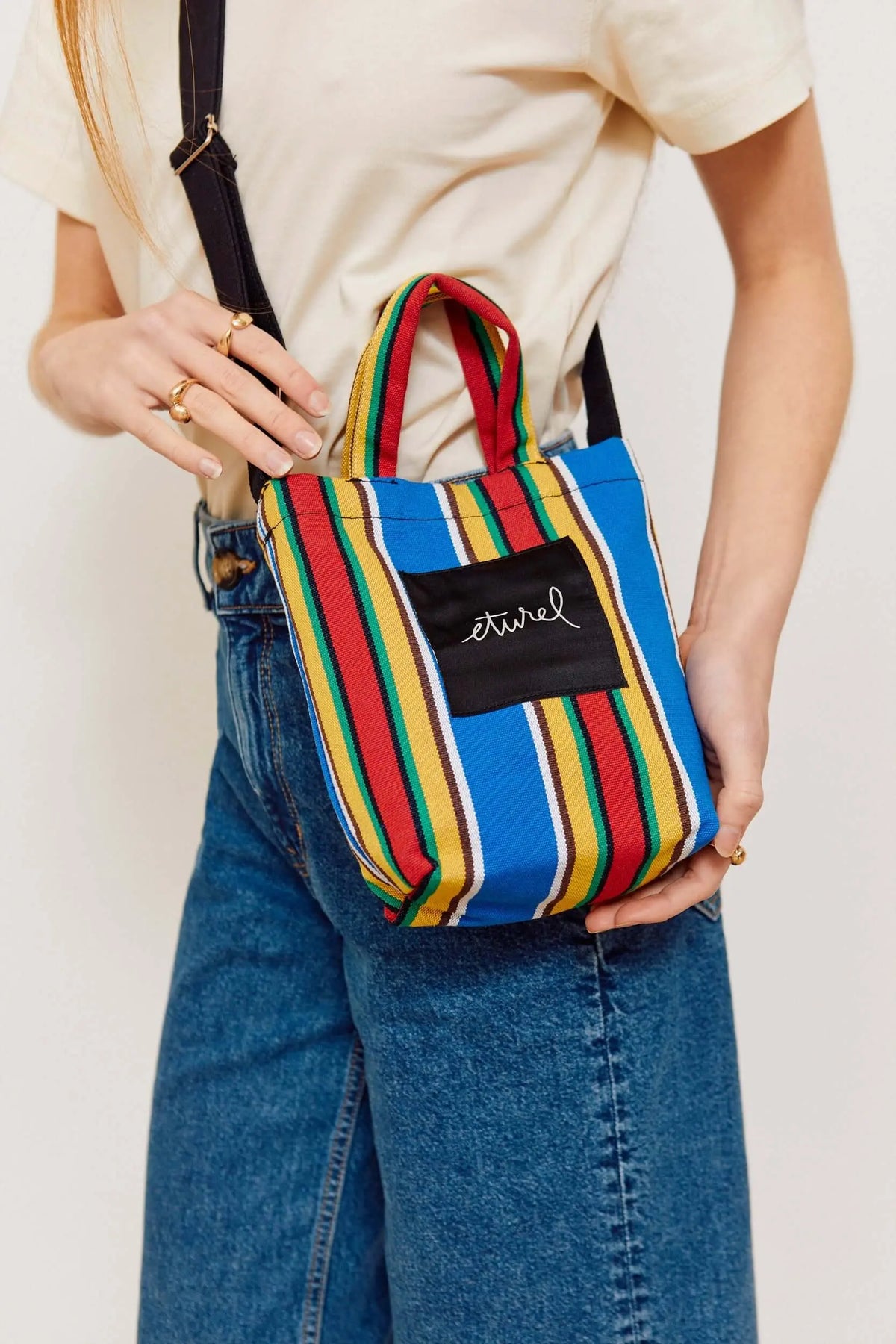 Bolso bandolera mini de lona con rayas multicolores, asas cortas y correa negra, usado por una persona.