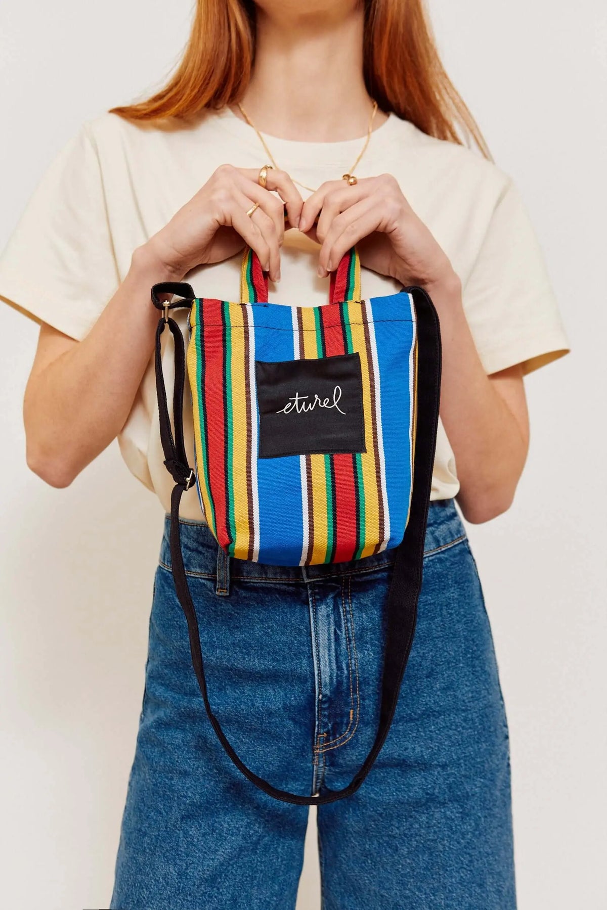 Mini bolso bandolera de lona a rayas multicolor con asas cortas, correa negra ajustable y estilo casual veraniego.