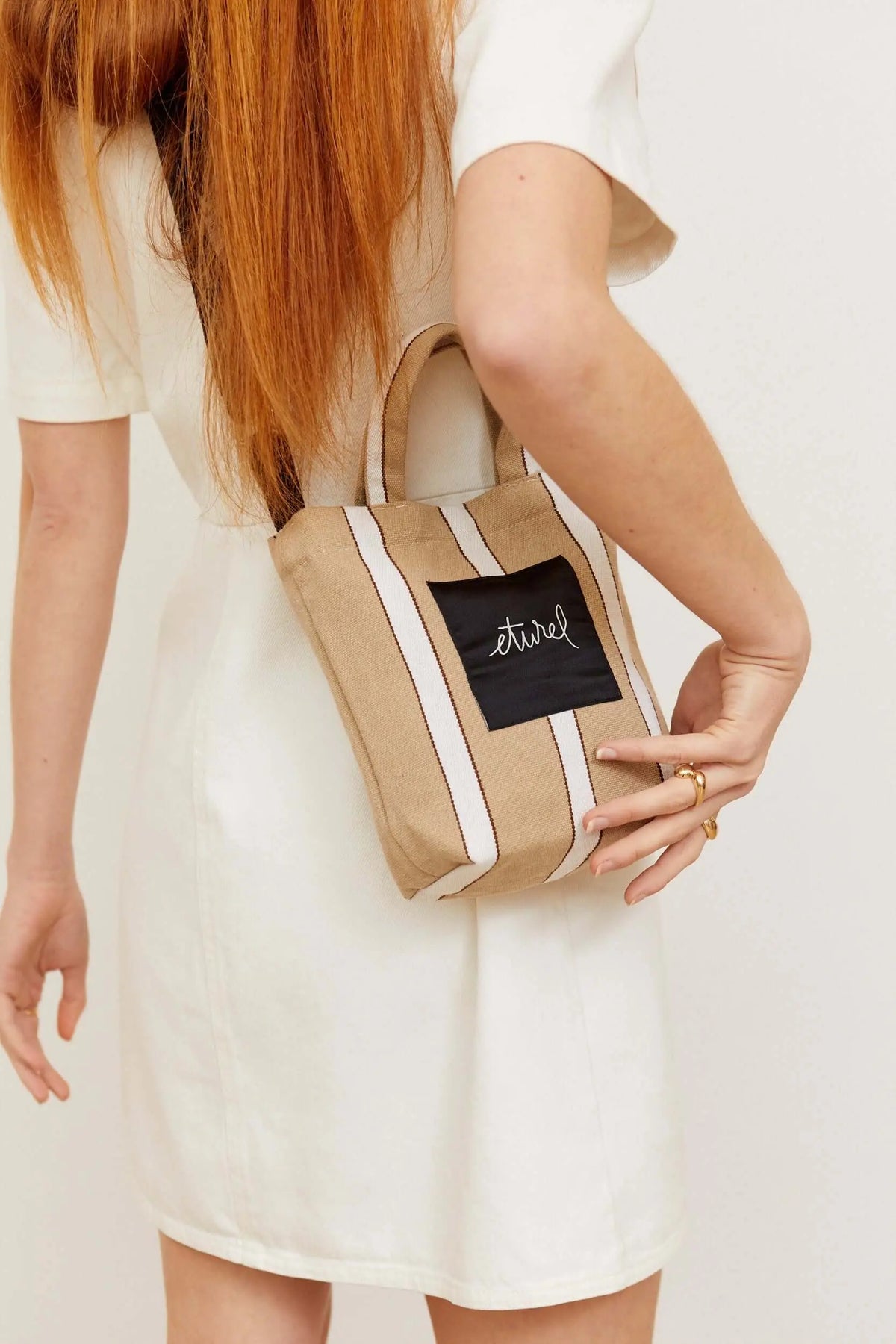 Bolso bandolera mini de lona beige con rayas blancas, asas cortas y parche central, estilo casual veraniego.