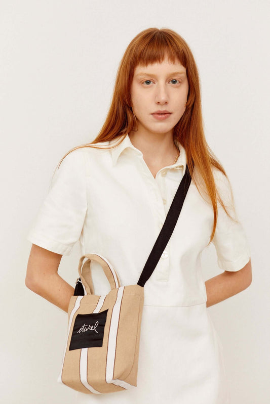 Mini beige canvas crossbody bag with white vertical stripes, top handles, and a black adjustable shoulder strap.