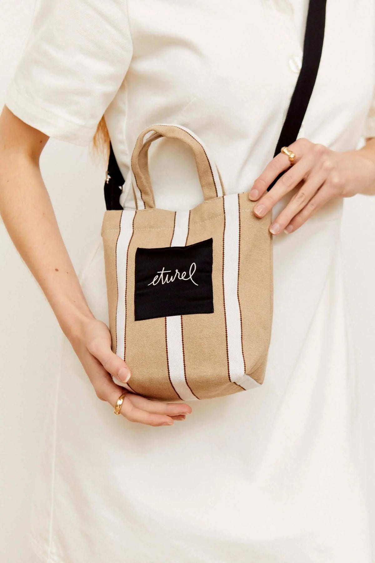Bolso bandolera mini de lona beige con rayas verticales blancas, asas cortas y correa negra ajustable.