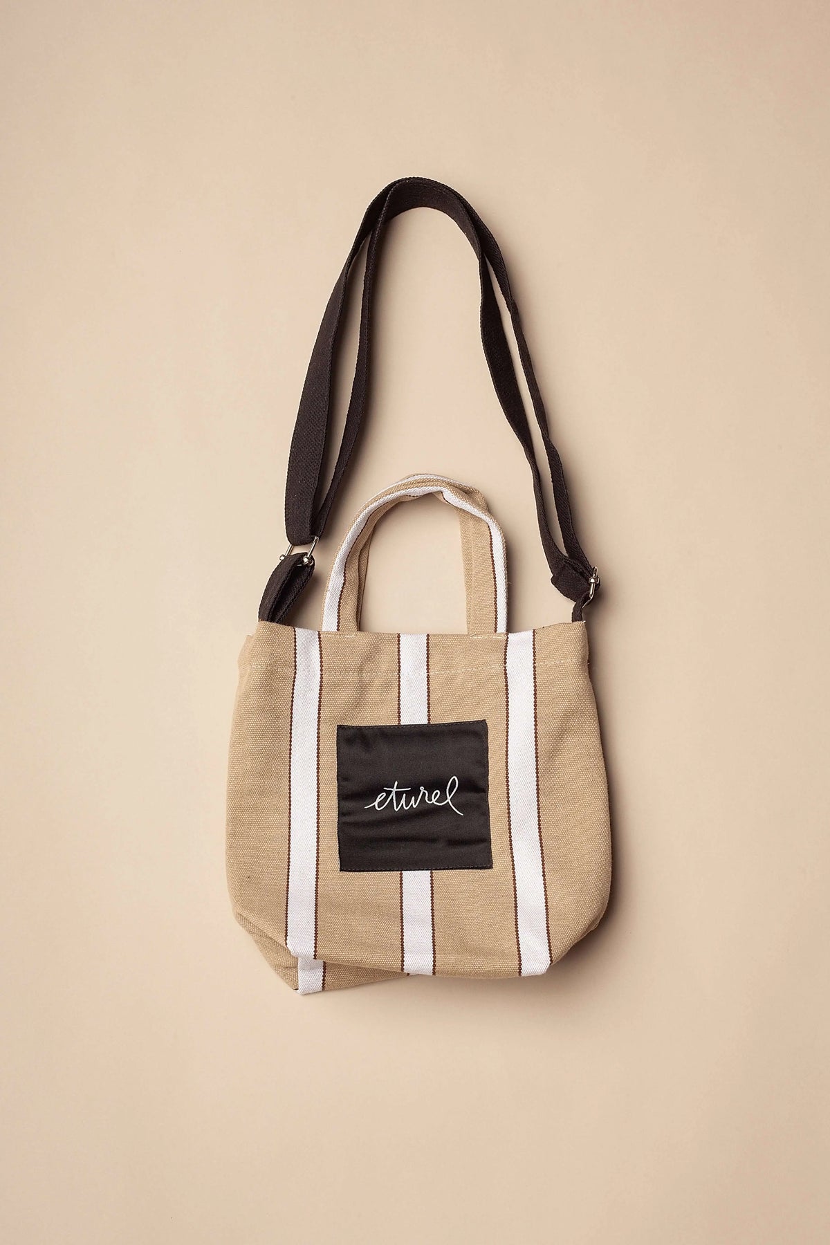 Beige canvas mini crossbody bag with white vertical stripes, top handles, and a black logo patch on the front.