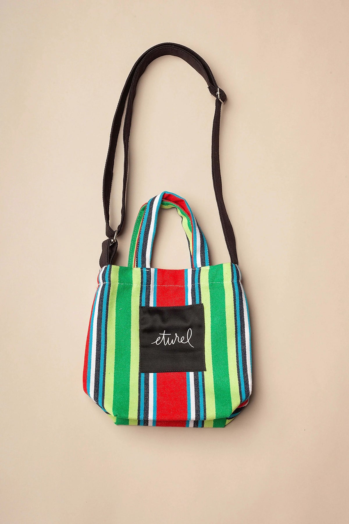 Bolso bandolera mini de lona a rayas multicolores con asas cortas, correa ajustable negra y bolsillo central frontal.