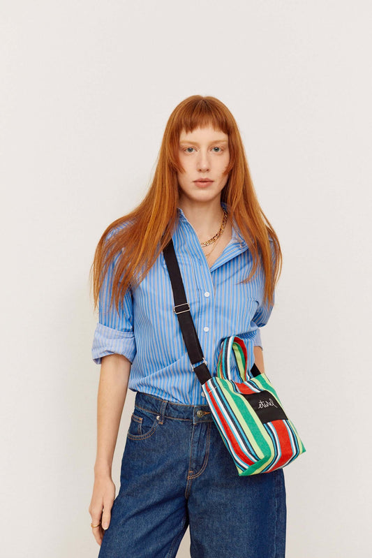 Bolso bandolera mini de lona a rayas multicolores con asa negra, estilo casual, llevado por una modelo.
