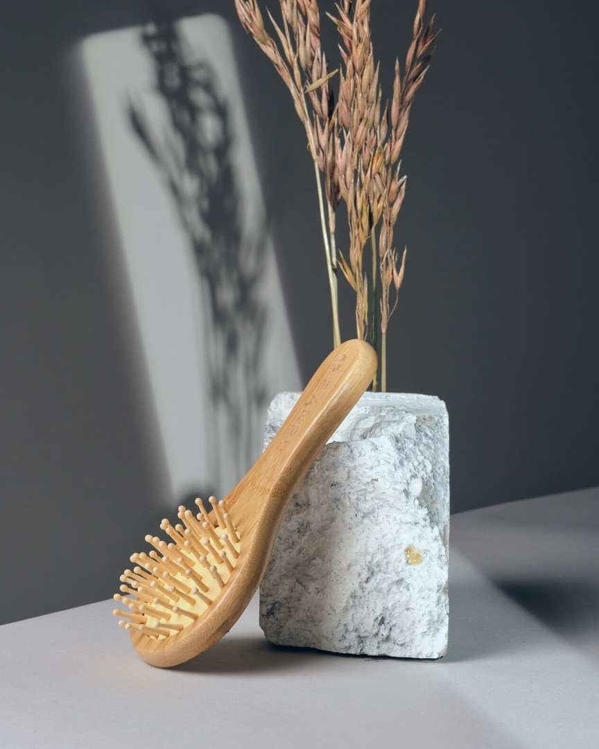 Cepillo de viaje Banbu de madera clara apoyado en una base de piedra blanca sobre un fondo gris con espigas secas.