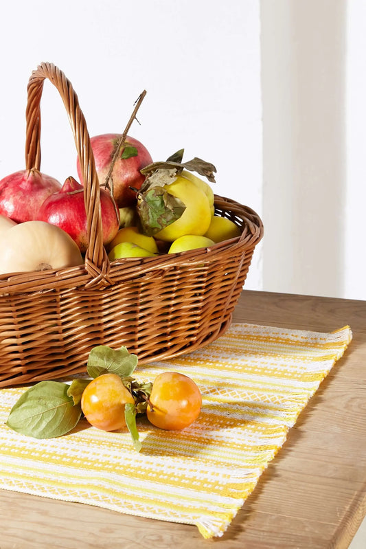 Camino de mesa de tela tejido en tonos amarillos y blancos con textura, sobre mesa de madera con cesta de fruta.