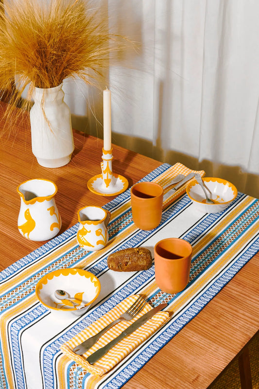 Juego de mesa colorido con cerámica blanca y amarilla, manteles de rayas azules y naranjas, y decoraciones modernas.