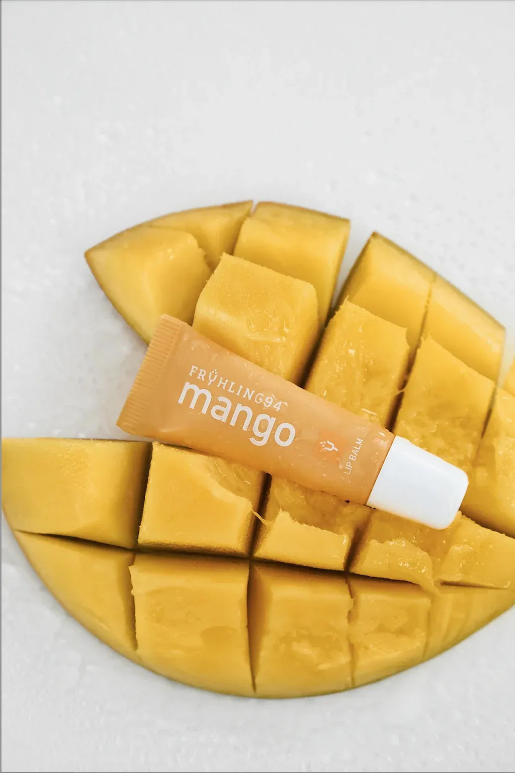 A squeeze tube of mango-scented lip balm labeled "FRÜHLING94" resting on a diced, juicy, yellow mango.