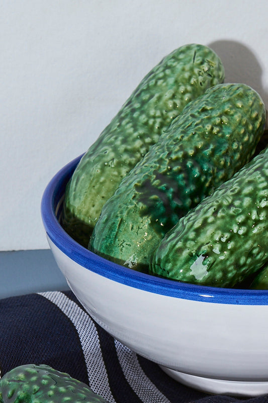 Cuenco cerámico blanco con borde azul, lleno de pepinos verdes brillantes. Ideal para decoración o cocina.