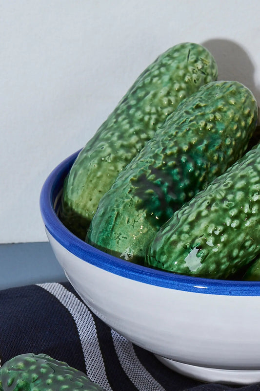 Cuenco cerámico blanco con borde azul, lleno de pepinos verdes brillantes. Ideal para decoración o cocina.