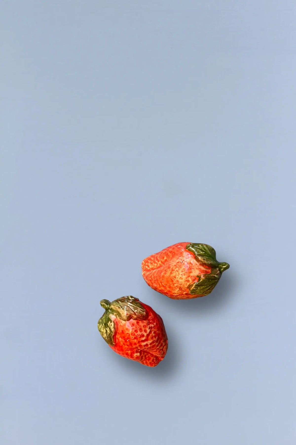 Fresas de cerámica roja brillante con hojas verdes sobre fondo azul suave. Ideal para decoración de cocina.