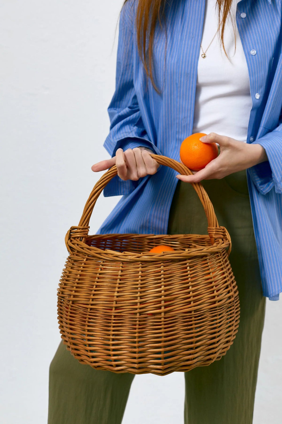 Cesta redonda de mimbre natural trenzado, color madera, con asa superior curva, sosteniendo naranjas en su interior.