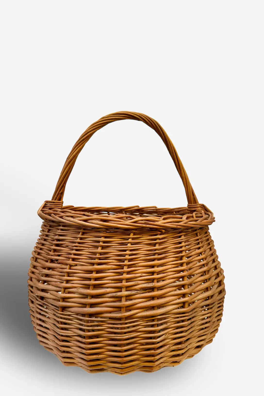 Cesta redonda de mimbre natural, color marrón claro, con tejido trenzado artesanal y asa superior arqueada.