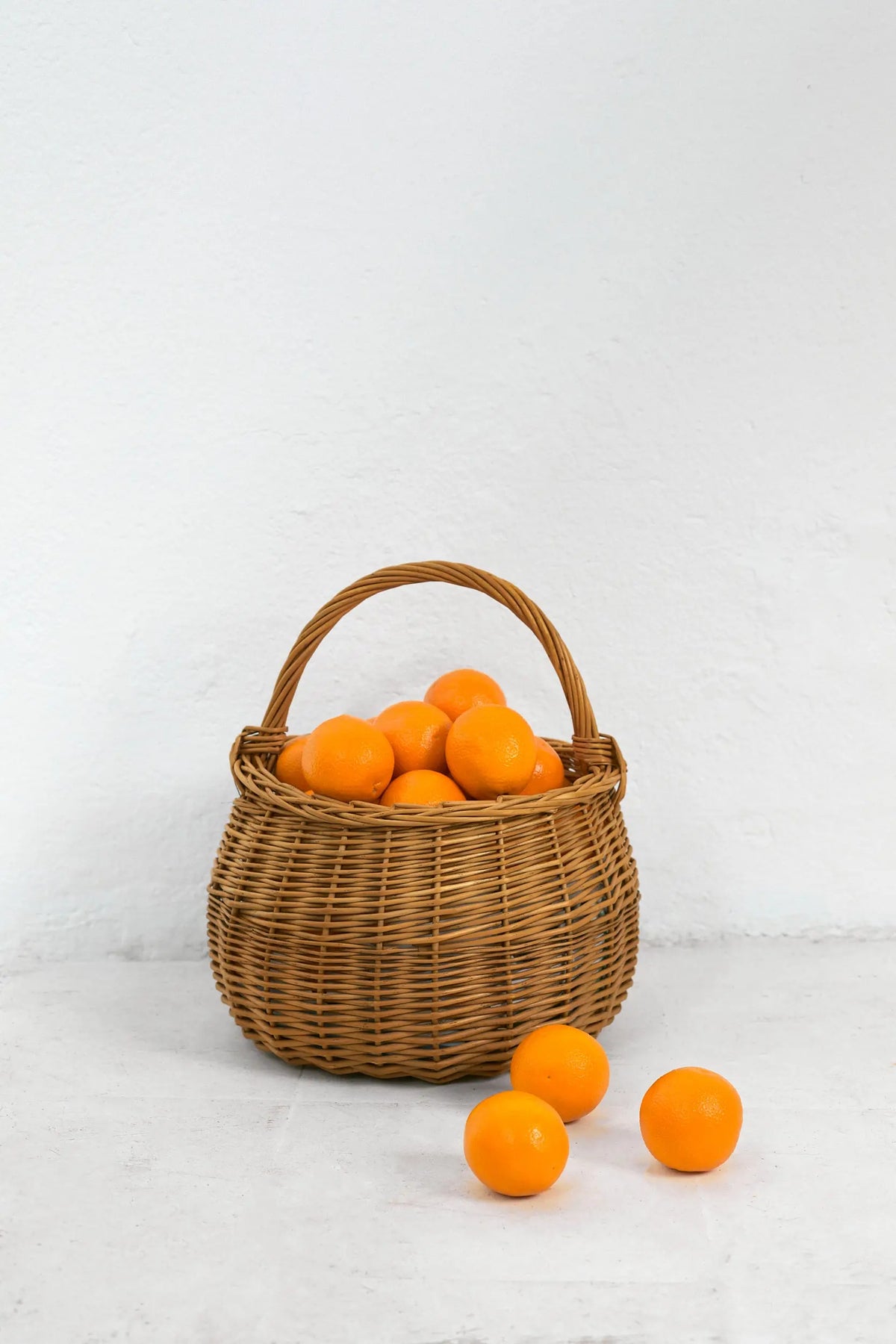 Cesta redonda de mimbre natural con asa, llena de naranjas frescas sobre un fondo blanco.