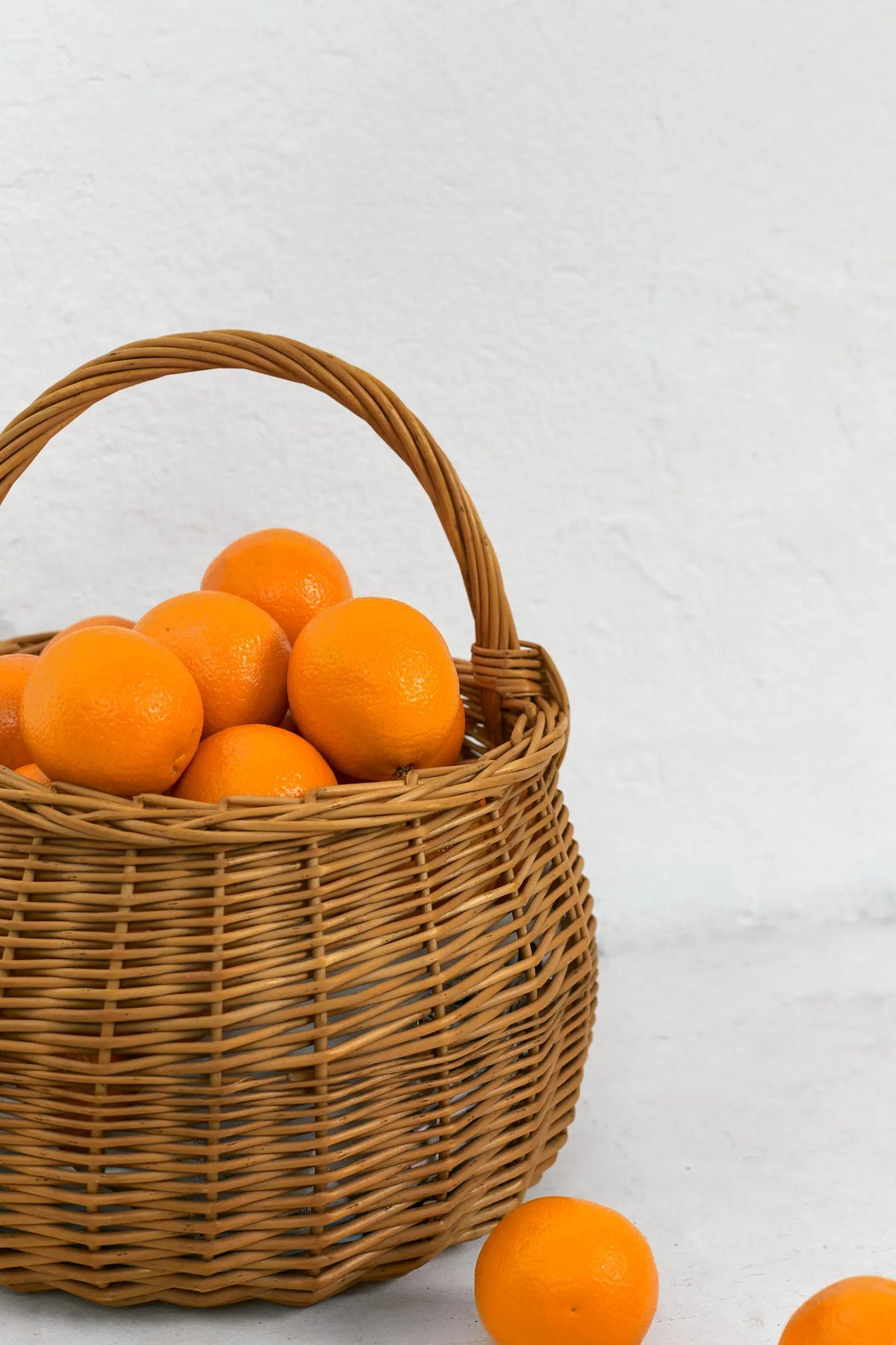 Cesta redonda de mimbre natural con asa, rellena de naranjas frescas sobre un fondo blanco.