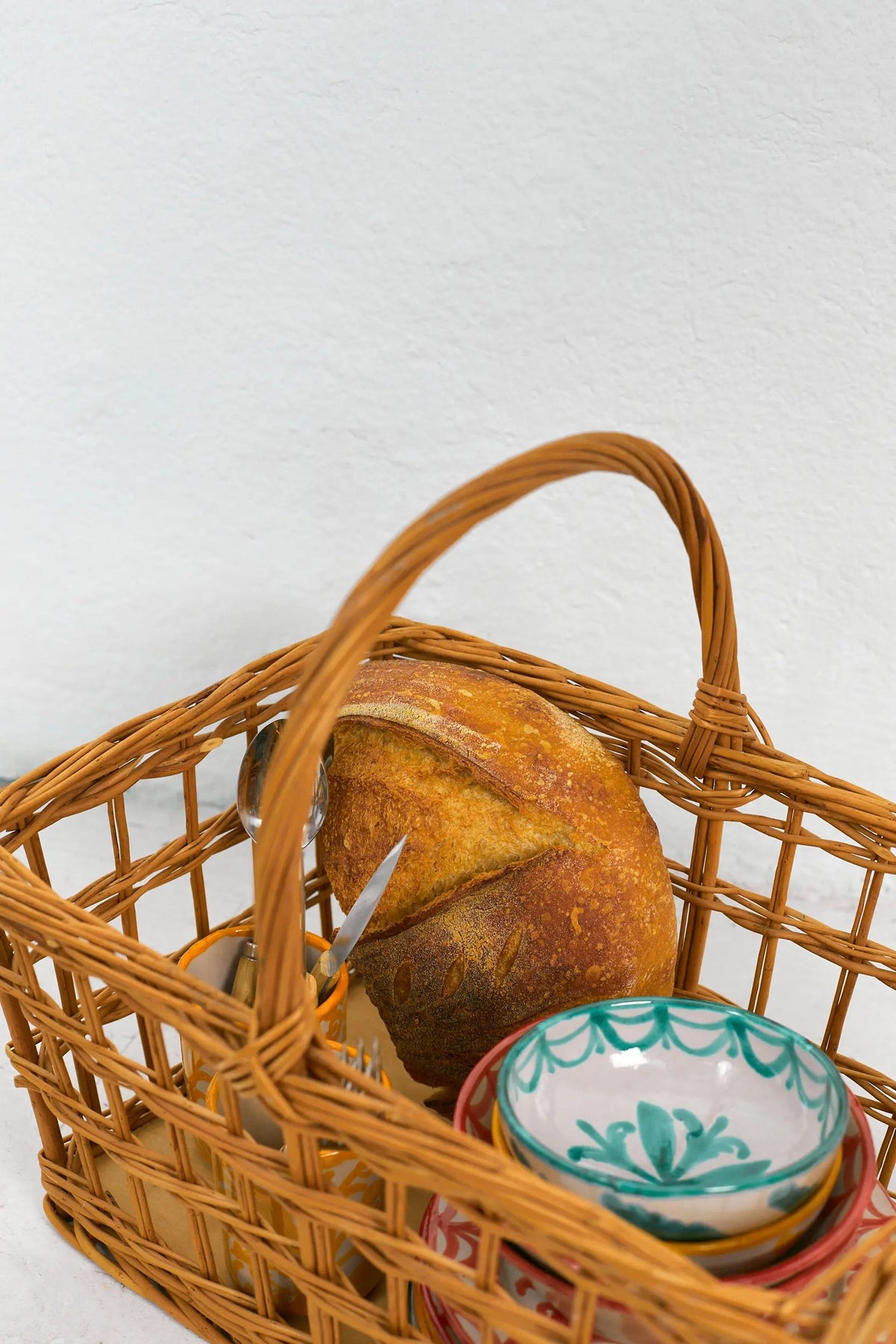 Natural wicker picnic basket with open-weave sides, containing a rustic bread loaf and decorative ceramic bowls.