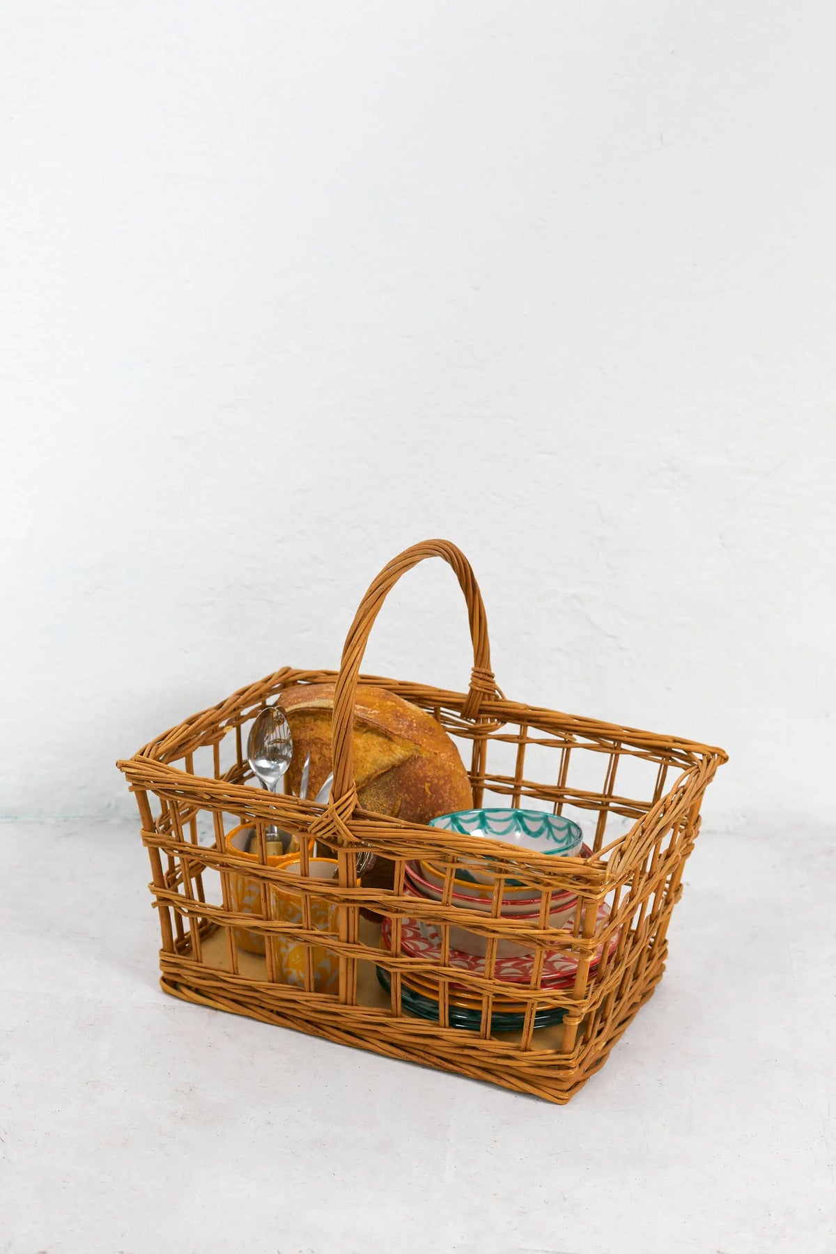 Rectangular open-weave natural wicker picnic basket with a high arched handle, containing bread and patterned plates.