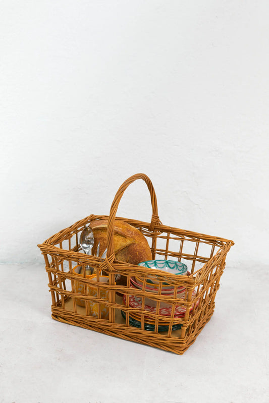 Rectangular open-weave natural wicker picnic basket with a high arched handle, containing bread and patterned plates.