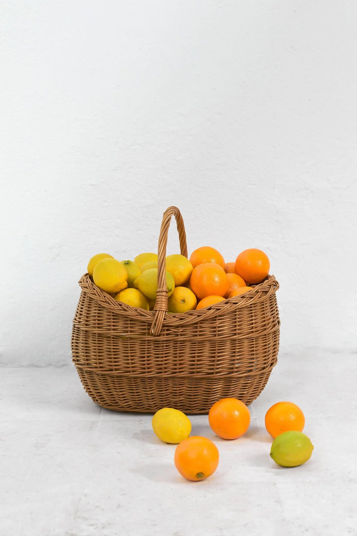 Cesta capazo de mimbre natural con asa central, llena de limones y naranjas frescas sobre fondo blanco.