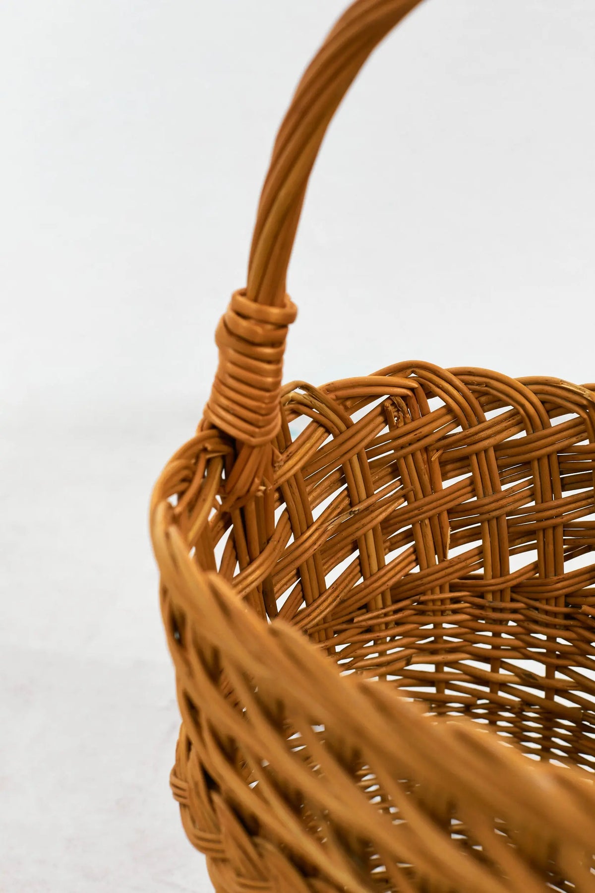 Cesta de mimbre natural trenzado, color marrón claro, con forma redondeada y asa arqueada fija. Fondo blanco.